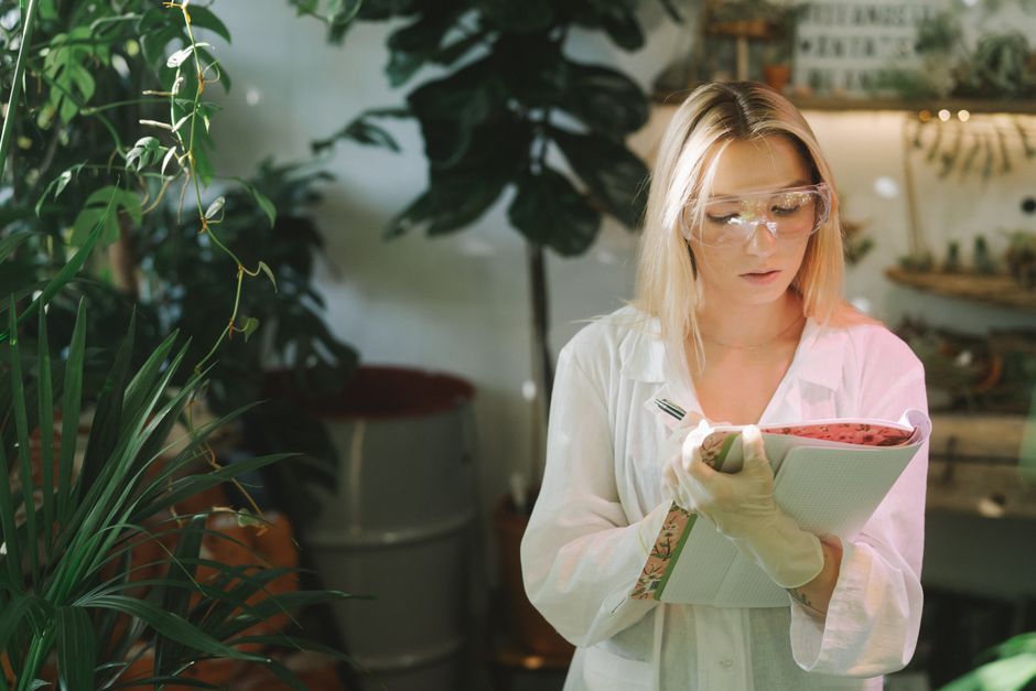 Focused female botanist writing down observations in a laboratory surrounded by plants.